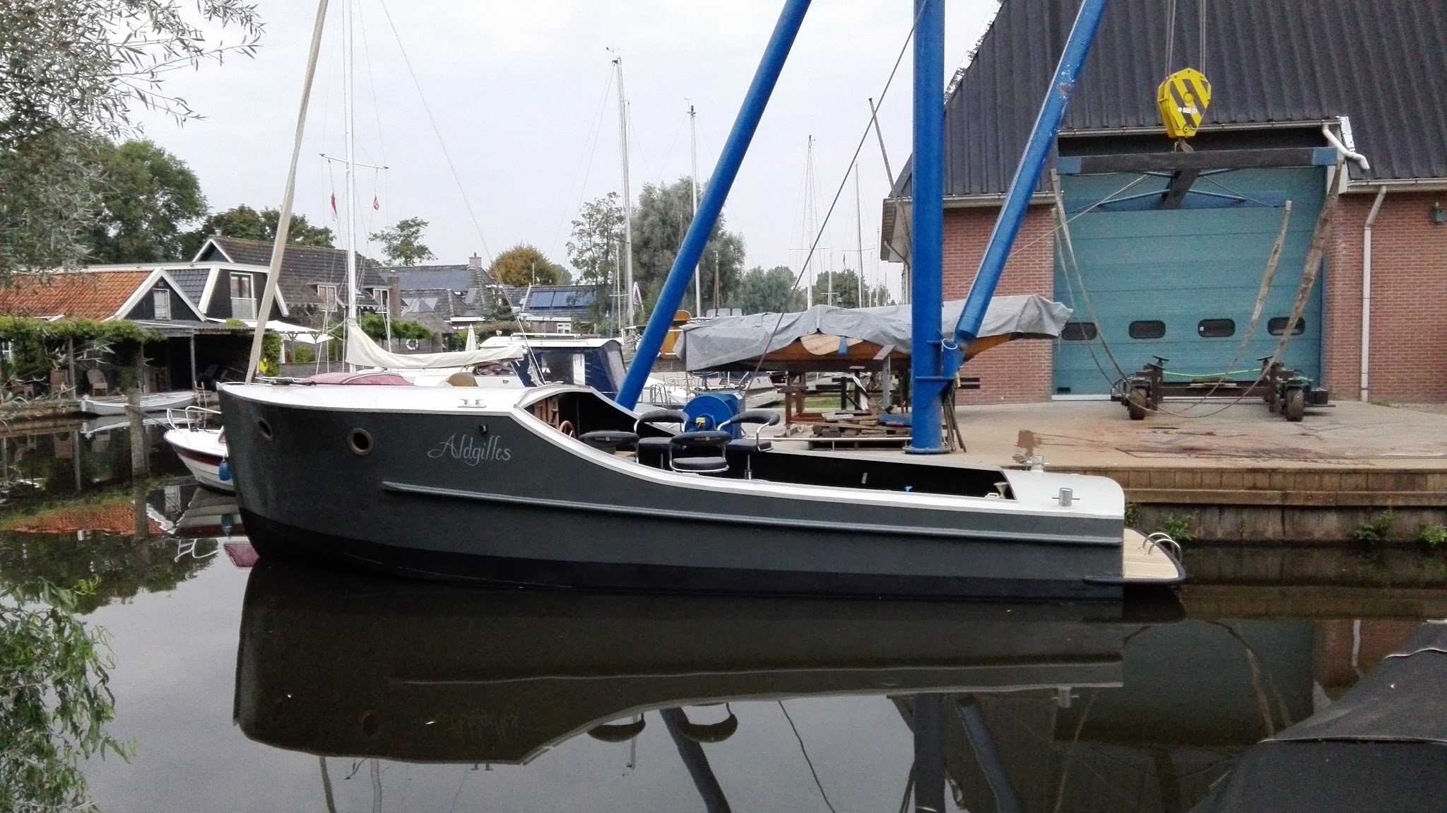 Een donkere sloep afgemeerd op spiegelglad water, met de blauwe kraan en de werkplaats erachter